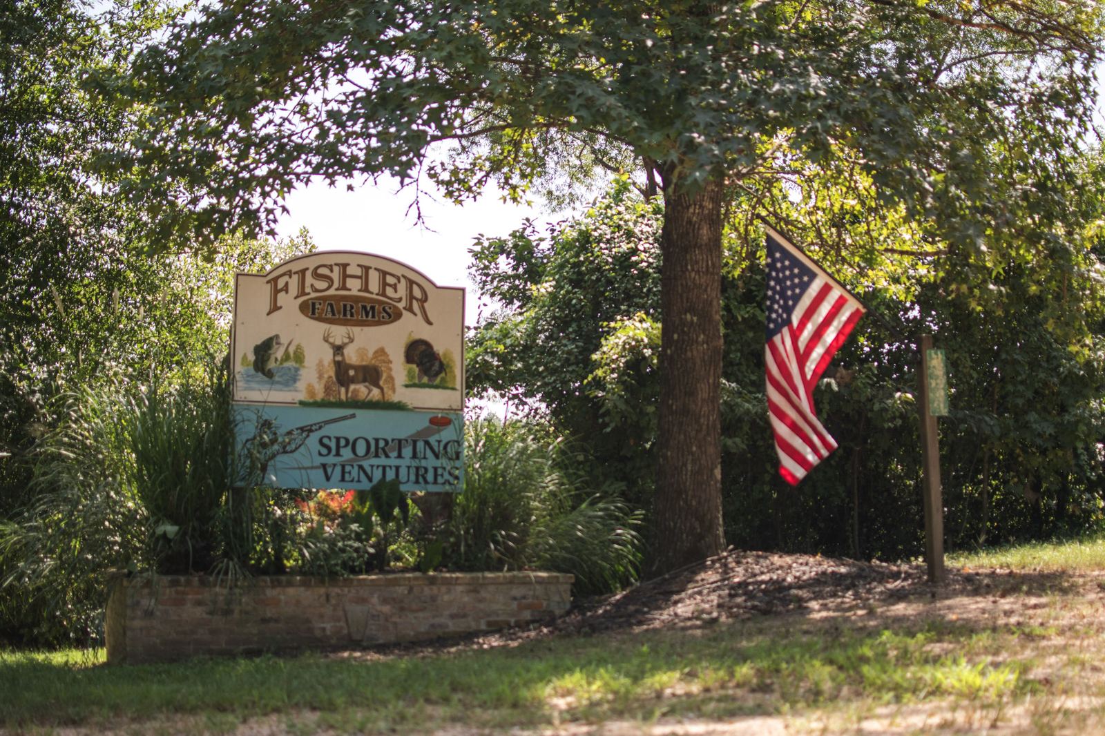 The Fisher Farms Sporting Ventures sign at the road, painted with a fish, a deer and a turkey above a shotgun and clay panel, with an American flag beside it